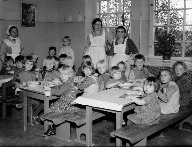 Kinder sitzen an Tischen in einem Klassenzimmer mit Tellern voller Essen, umgeben von Bänken, einem Foto auf der Wand und Fenstern mit Pflanzen draußen.