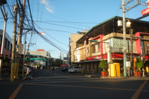 Eine Stadtstraße mit Autos, Fußgängern, Strommasten, Verkehrsampeln, Gebäuden mit Schildern, Topfpflanzen und einem bewölkten Himmel.