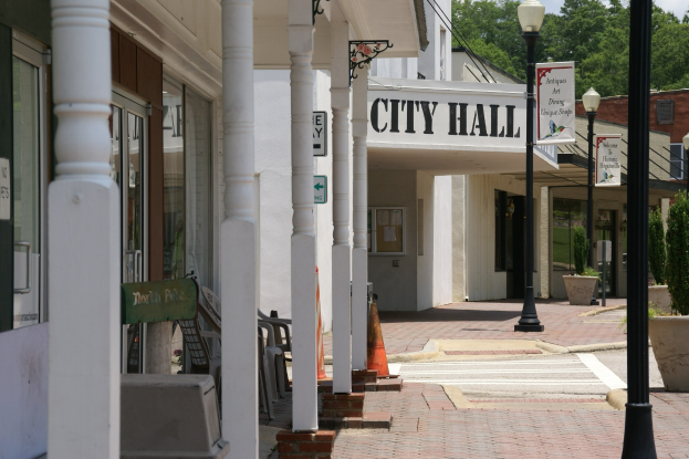 Rathausgebäude mit umliegender Infrastruktur, darunter Straßenmasten, Lichter, Namenschilder, Fahrzeuge, Verkehrskegel, Stühle, Topfpflanzen, Bäume und eine bewölkte Himmel.