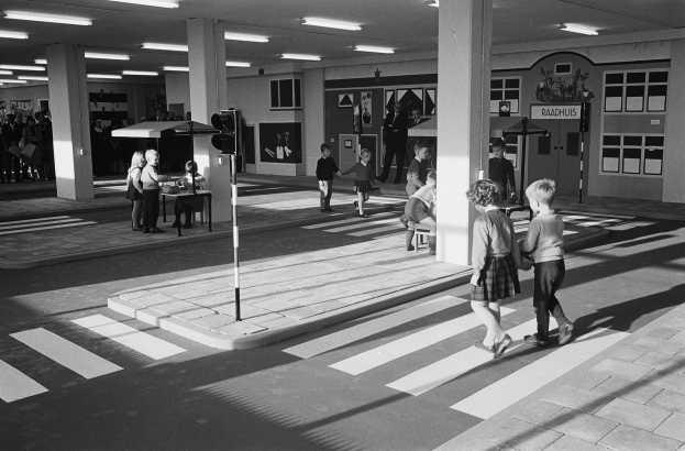 Schwarzes und weißes Foto von Kindern, die eine Straße an einer Ampel überqueren, mit Fußgängern in der Nähe, einem Verkehrssignalmast im Vordergrund und Säulen mit Deckenleuchten im Hintergrund.