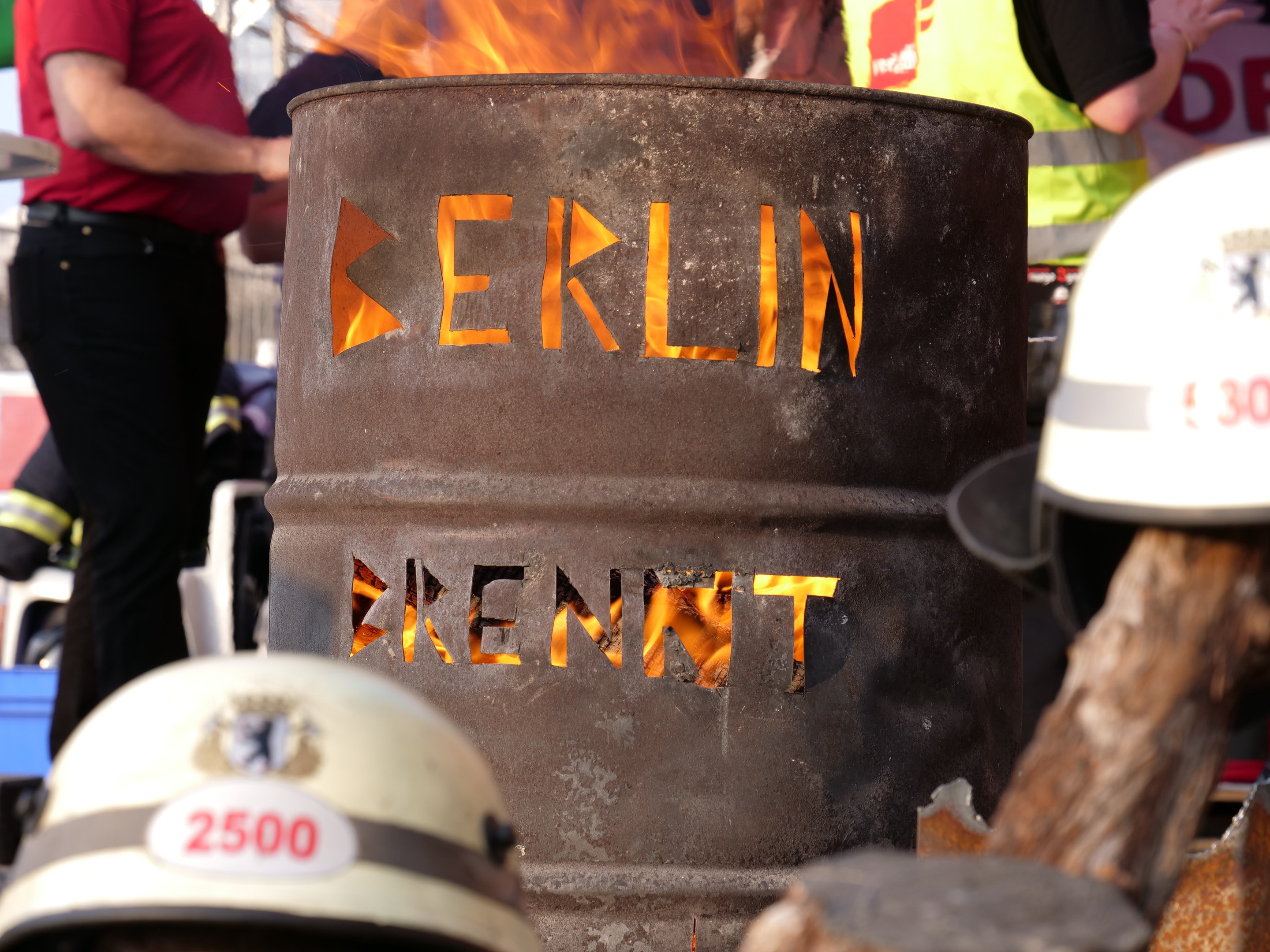 Menschen um ein Feuerhydrant mit der Aufschrift "Berlin" mit einem brennenden Fässer im Vordergrund, Helme unten und ein hölzernes Objekt rechts.