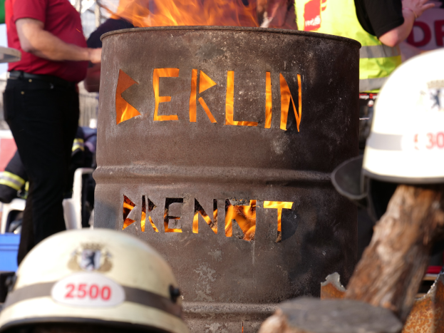 Menschen um ein Feuerhydrant mit der Aufschrift "Berlin" mit einem brennenden Fässer im Vordergrund, Helme unten und ein hölzernes Objekt rechts.