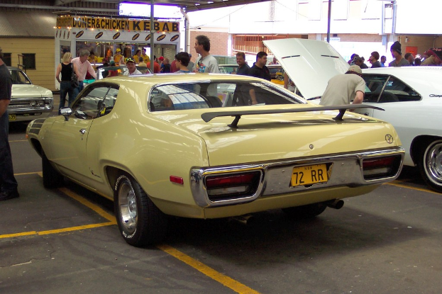 Ein Oldtimer fährt mit Menschen in der Nähe, ein Geschäft in der Mitte und Gebäuden im Hintergrund.