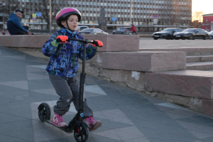 Ein junger Junge fährt mit einem Helm und Handschuhen auf einem Scooter einen Gehweg entlang, mit Stufen, Fahrzeugen, Menschen, Bäumen, Pfosten, Brettern, Gebäuden und einem klaren blauen Himmel im Hintergrund.