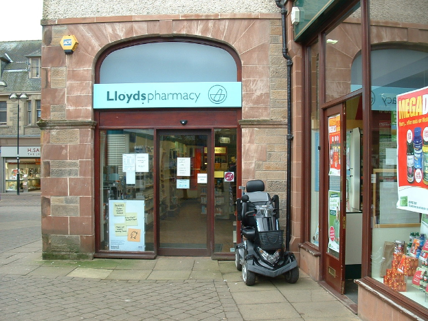 Eine Apotheke mit einem vor ihr geparkten Fahrzeug und einem Gebäude, das links sichtbar ist.