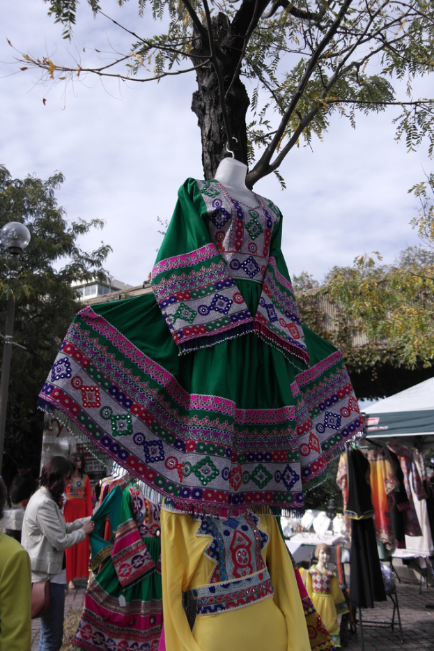 Eine Schaufensterpuppe steht in der Mitte eines belebten Einkaufsgebiets mit mehreren Geschäften, eine Person, die Kleider an einem Ständer arrangiert, ein Gebäude und Bäume im Hintergrund.