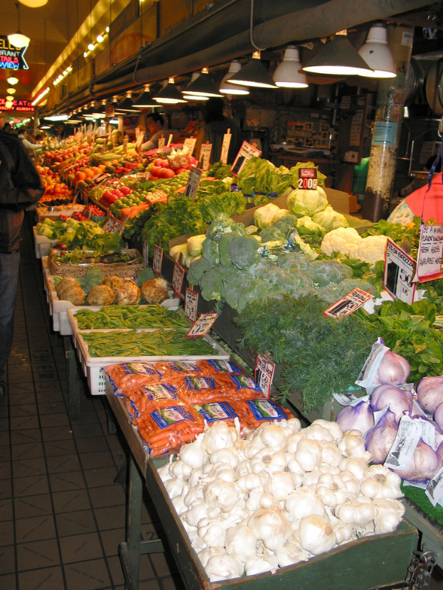 Eine MarktSzene mit zahlreichen Körben, gefüllt mit verschiedenen Gemüsen und Früchten, einschließlich Knoblauch, Blattgemüse, Kohlsorten und Brokkoli, mit Menschen im Hintergrund und Preisschildern auf den Körben, unter Deckenbeleuchtung.