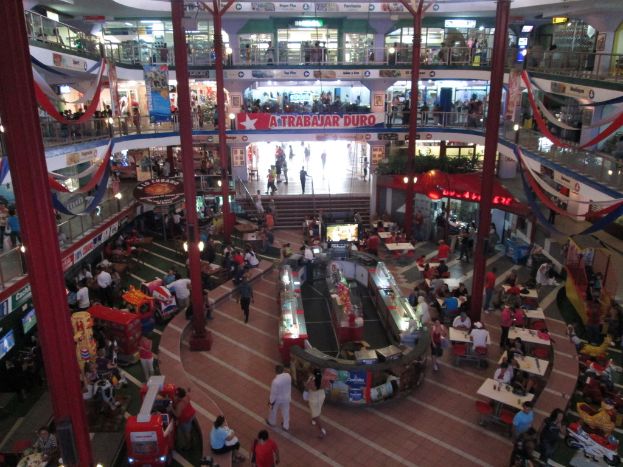 Eine belebte Einkaufszeile mit Säulen, Menschen, Tischen, Fliesen, Flaggen, Geschäften, einem Banner, Treppen und einem Namensschild.