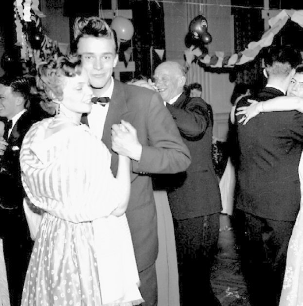 Ein Schwarz-Weiß-Foto eines Mannes und einer Frau, die auf einer Party tanzen, umgeben von lachenden Menschen und Ballons im Hintergrund.