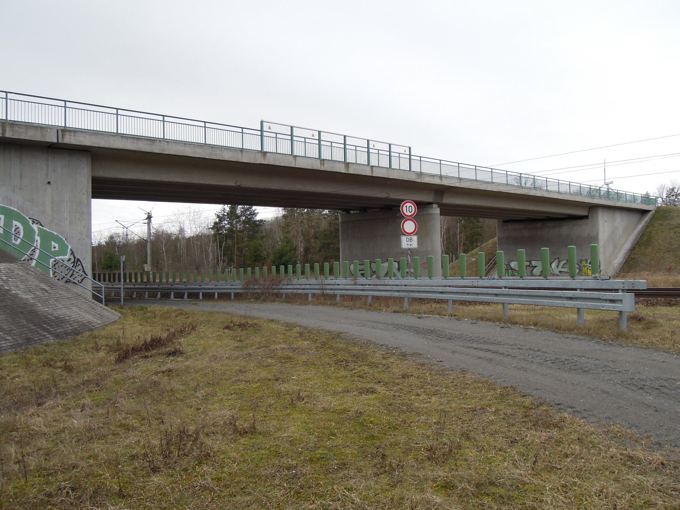 Brücke über einen Schotterweg mit Graffiti-beschmierten Geländern, Pfählen, Schildern, einem Zaun, Gras, Bäumen und Himmel im Hintergrund.