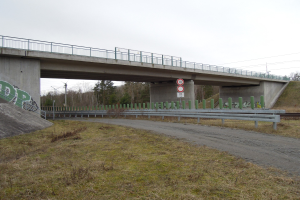 Brücke über einen Schotterweg mit Graffiti-beschmierten Geländern, Pfählen, Schildern, einem Zaun, Gras, Bäumen und Himmel im Hintergrund.