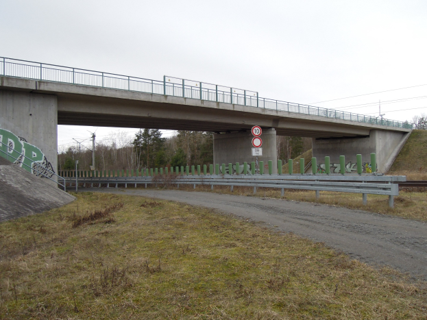 Brücke über einen Schotterweg mit Graffiti-beschmierten Geländern, Pfählen, Schildern, einem Zaun, Gras, Bäumen und Himmel im Hintergrund.
