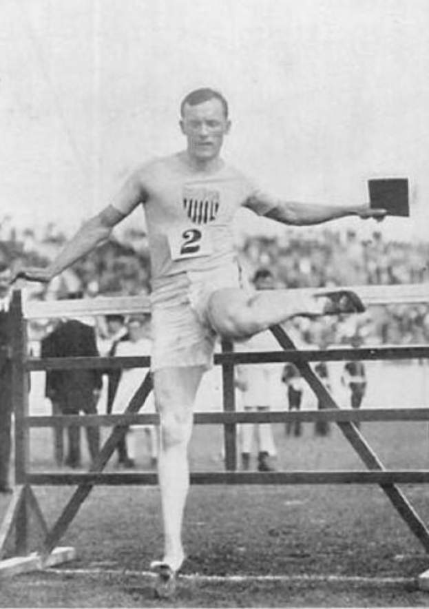 Ein Schwarz-Weiß-Foto eines Mannes in Sportkleidung, der über ein Hindernis springt, mit Zuschauern im Hintergrund und dem Himmel darüber.