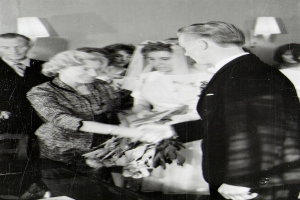 Ein Schwarz-Weiß-Foto eines Brautpaares beim Austausch der Eheringe, wobei die Braut ein weißes Kleid und eine Schleppe trägt und einen Strauß hält, und der Bräutigam einen Anzug und eine Krawatte trägt, mit Wandlampen im Hintergrund.