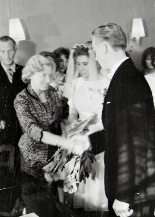 Ein Schwarz-Weiß-Foto eines Brautpaares beim Austausch der Eheringe, wobei die Braut ein weißes Kleid und eine Schleppe trägt und einen Strauß hält, und der Bräutigam einen Anzug und eine Krawatte trägt, mit Wandlampen im Hintergrund.