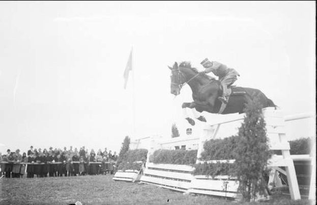 Schwarz-weißes Foto eines Pferdes und seines Reiters, die über ein Hindernis springen, bei den 1953 Royal Ascot Horse Trials, mit Zuschauern auf der linken Seite, einer Fahne im Hintergrund und Gras am Boden.