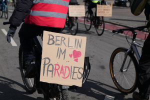 Eine Gruppe von Menschen, die auf Fahrrädern eine Straße entlangfährt, mit einem "Berlin I'm Paradies"-Schild im Vordergrund und einem unscharfen Auto im Hintergrund.