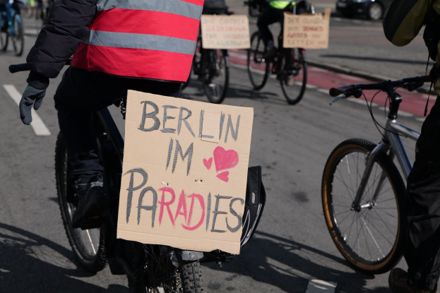 Eine Gruppe von Menschen, die auf Fahrrädern eine Straße entlangfährt, mit einem "Berlin I'm Paradies"-Schild im Vordergrund und einem unscharfen Auto im Hintergrund.