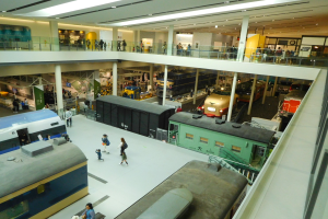 Ein großer Museumsraum mit verschiedenen Zugtypen, mit Menschen, die beobachten, Geländern, Säulen, Deckenleuchten und Informationsschildern.