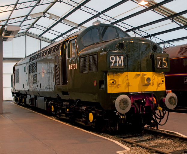 Ein Zug fÃ¤hrt auf den Schienen neben einem Bahnsteig mit einem Mast auf dem Dach, der MetallstÃ¤be mit Lichtern trÃ¤gt.