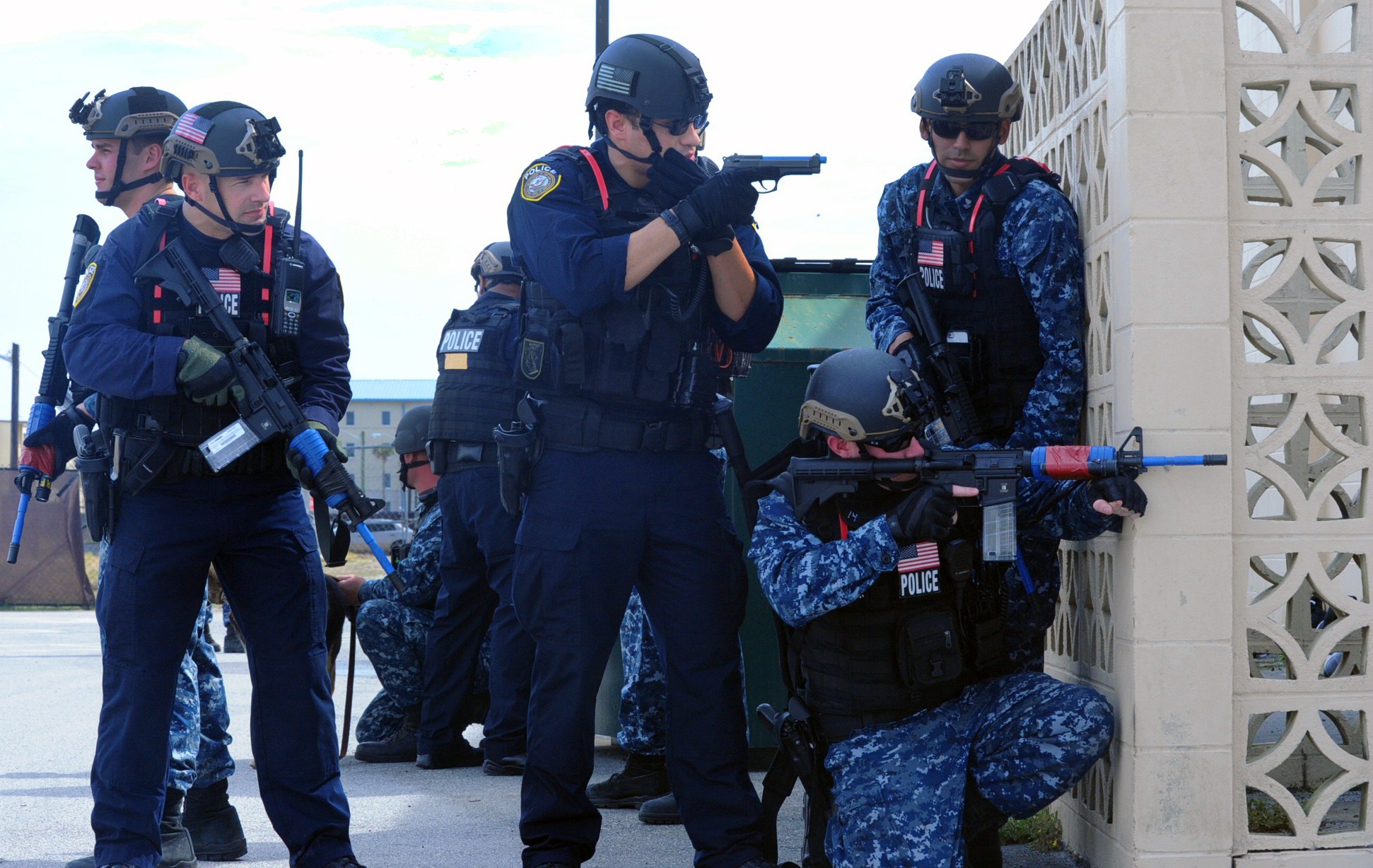 Gruppe von Polizisten in Helmen und Uniformen auf einer Brücke stehend, Waffen haltend, mit einer Wand auf der rechten Seite und Gebäuden, Bäumen und Himmel im Hintergrund.