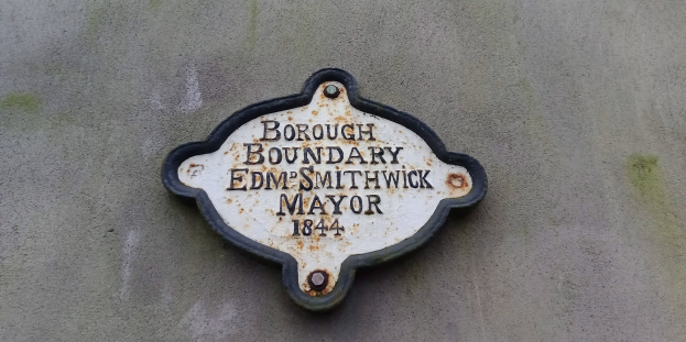 Metallplatte an der Außenseite eines Gebäudes, die die Borough-Grenze markiert, mit "Borough Boundary Edm Smithwick Mayor" in fetten weißen Buchstaben auf einem schwarzen Hintergrund.