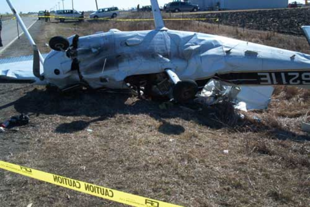 Wrack eines Kleinflugzeugs, das auf der Seite einer Straße abgestürzt ist, umgeben von Gras und Absperrband, mit Fahrzeugen, Polen, Bäumen und einem klaren blauen Himmel im Hintergrund.