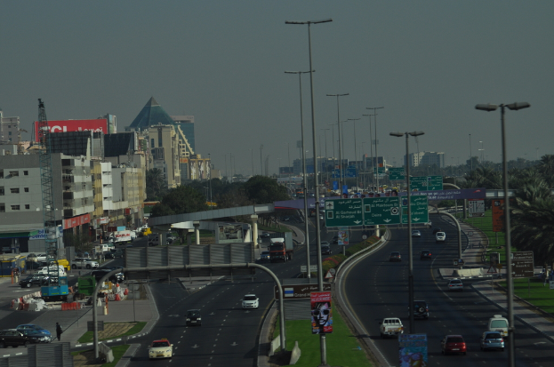 Eine Luftaufnahme einer Stadt mit Gebäuden, Türmen, Kraftfahrzeugen, Werbetafeln, Schildern, Straßen, Bäumen, Straßenlaternen, Menschen, Verkehrsleitkegeln und Informationsanzeigen unter einem klaren Himmel.