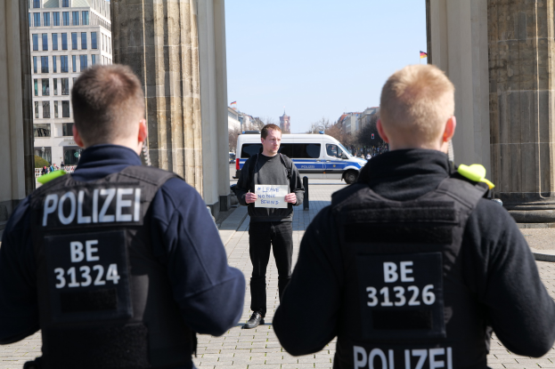 Drei Polizisten stehen vor einem Gebäude, ein Mann in der Mitte hält ein Papier, flankiert von Säulen; Fahrzeuge, Menschen, Bäume und Gebäude sind im Hintergrund zu sehen.
