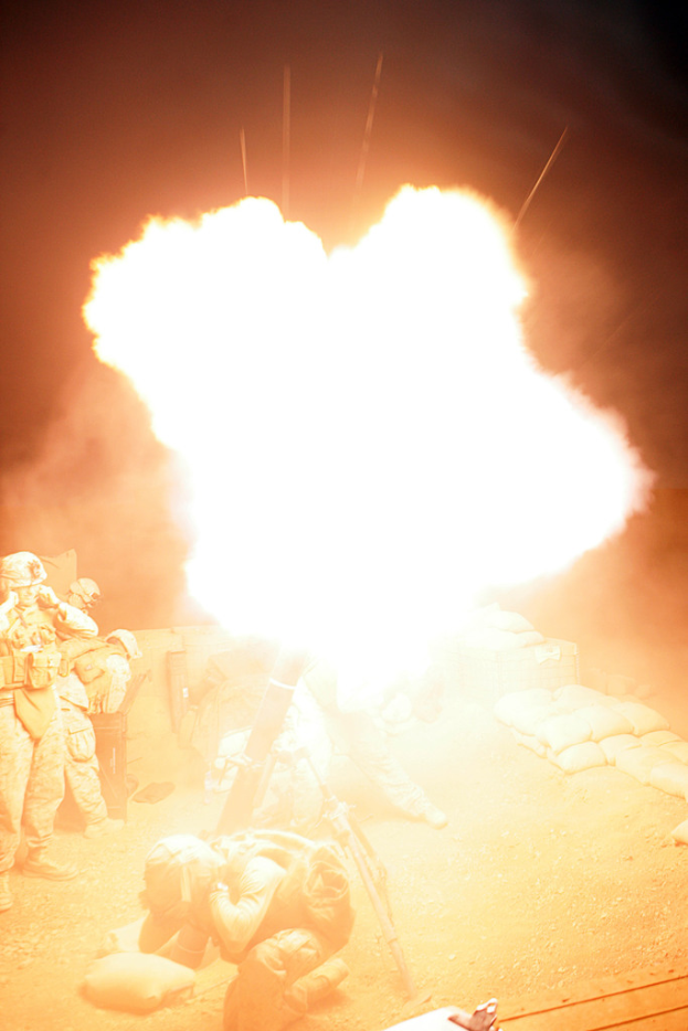 Mehrere Personen in Uniformen und Helmen auf einem Gelände mit ein paar Säcken herumliegen, mit einem Feuer in der Mitte der Szene.