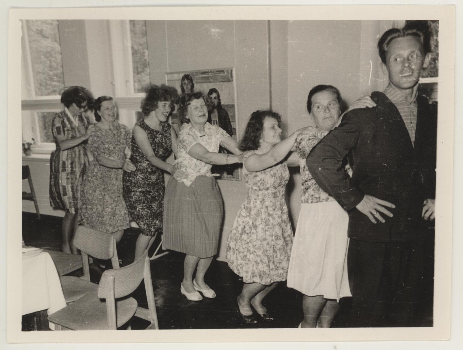 Eine Gruppe von Menschen, die in einem Raum mit Stühlen und einem mit wei├čer Decke bedeckten Tisch tanzen. An der Wand h├Ąngt ein Foto und im Hintergrund sind Fenster zu sehen.