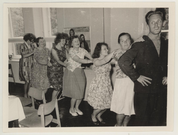 Eine Gruppe von Menschen, die in einem Raum mit Stühlen und einem mit wei├čer Decke bedeckten Tisch tanzen. An der Wand h├Ąngt ein Foto und im Hintergrund sind Fenster zu sehen.
