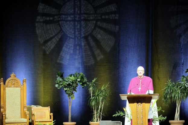 Ein Mann in einem pink-weißen Kleid steht neben einem Rednerpult in einem Auditorium, mit Pflanzen und zwei Stühlen daneben und einer bunten Wand im Hintergrund.
