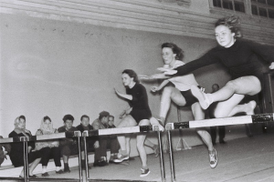Zwei Frauen in der Luft beim Überqueren eines Hindernisses in einem Gymnastikraum, mit Zuschauern links und Fenstern an der Rückwand.
