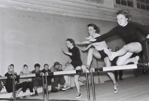 Zwei Frauen in der Luft beim Überqueren eines Hindernisses in einem Gymnastikraum, mit Zuschauern links und Fenstern an der Rückwand.