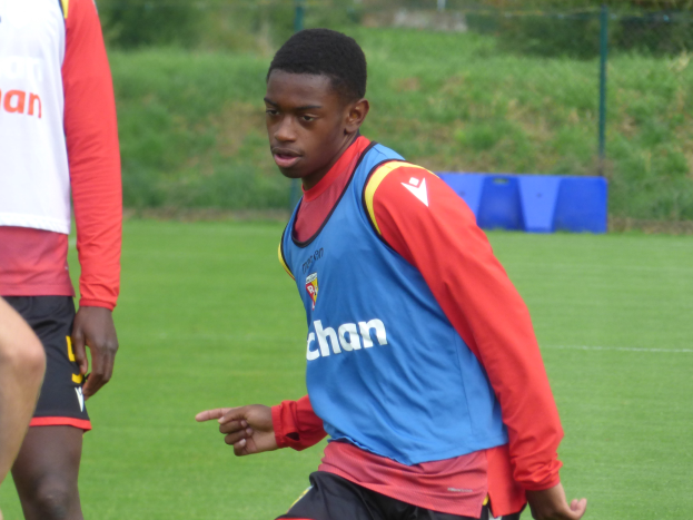Ein junger Mann in einem blauen Hemd und schwarzen Shorts spielt Fußball auf einem grünen Feld, umgeben von einem Zaun, Pflanzen und Bäumen und scheint mitten in einem Training zu sein.