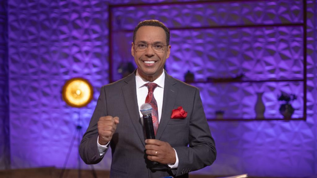 Stephen Colbert, Moderator von 'The Daily Show with Trevor Noah', steht auf einer Bühne und hält ein Mikrofon in der Hand, trägt einen Anzug, eine Krawatte und eine Brille, während im Hintergrund ein Scheinwerfer und ein Ständer zu sehen sind.