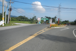 Eine vielbefahrene Hauptstraße mit Fahrzeugen, elektrischer Infrastruktur, Beschilderung und natürlichen Elementen wie Gras, Bäumen und Hügeln unter einem bewölkten Himmel.