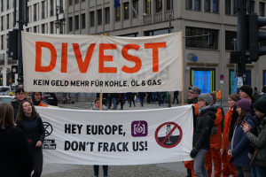 Eine Gruppe von Menschen mit einem Banner "Divest Hey Europe, Don't Frack Us" auf einer Straße mit Gebäuden, Straßeninfrastruktur, Fahrzeugen, Bäumen und einem klaren Himmel im Hintergrund.