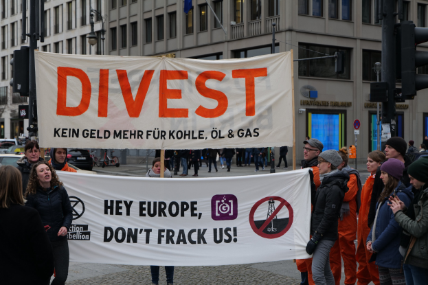 Eine Gruppe von Menschen mit einem Banner "Divest Hey Europe, Don't Frack Us" auf einer Straße mit Gebäuden, Straßeninfrastruktur, Fahrzeugen, Bäumen und einem klaren Himmel im Hintergrund.