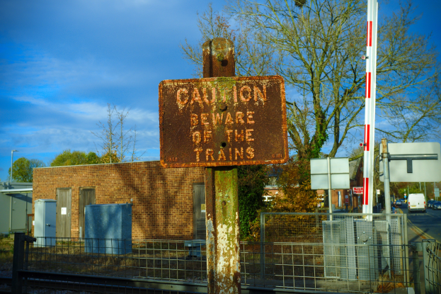Vorsichtshinweis auf einem Bahngleis neben einem Metallzaun, mit Bäumen, Strommasten, einem Gebäude, Containern, Fahrzeugen auf einer Straße und einem bewölkten Himmel.