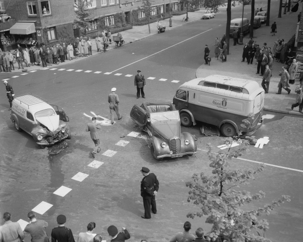 Schwarz-weiß-Szene eines Autounfalls an einer Straßenkreuzung mit einem Lieferwagen, anderen Fahrzeugen und einer Gruppe von Schaulustigen vor Häusern, Bäumen, Strommasten und Schildern.