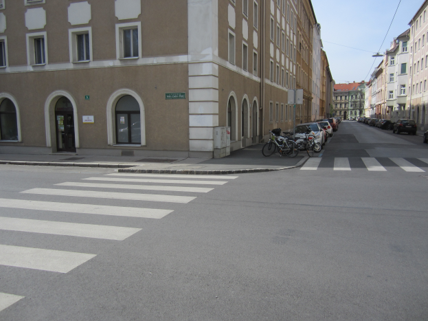 Eine Stadtstraße mit Zebrastreifen, Gebäude mit Fenstern und Türen, geparkte Fahrräder und ein klarer blauer Himmel.