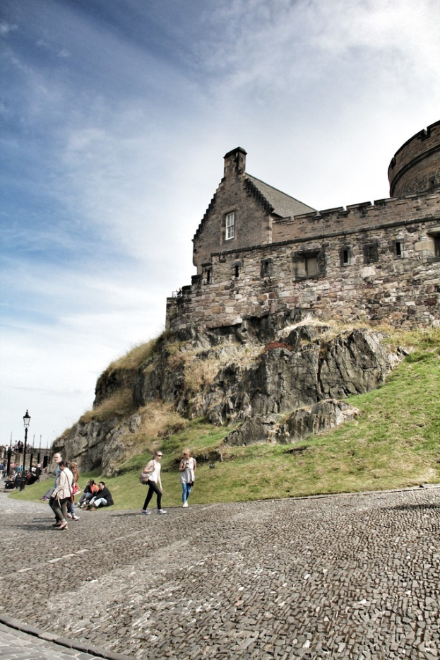 Ein Schloss auf einem Hügel mit Menschen auf dem Gehweg im Vordergrund, Bäume und Straßenlaternen links und Gras rechts, unter einem teilweise bewölkten, sonnigen Himmel.