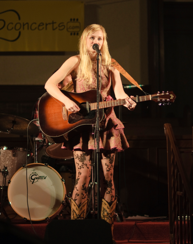 Eine Frau spielt Gitarre und singt auf der Bühne bei einem Konzert, mit einer Band und einem Bildschirm im Hintergrund und einer Bühne unten.
