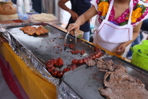 Eine Frau in einer Schürze kocht auf einem Grill an einem Markt, mit einem Tisch voller Lebensmittel, einem Eimer und anderen Gegenständen in der Nähe und ein paar Menschen im Hintergrund.
