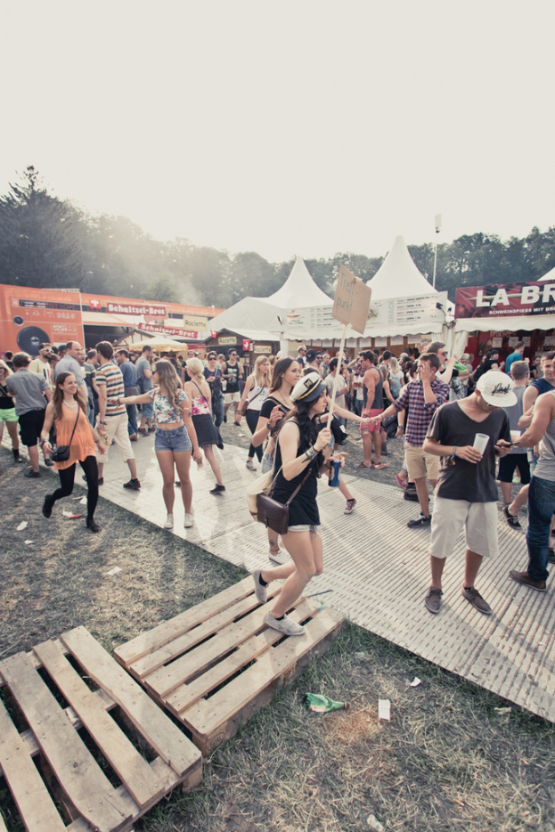 Menschen stehen und gehen auf einer Straße mit Zelten, Bäumen und Himmel im Hintergrund.