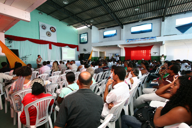 Eine Gruppe von Menschen, die auf Stühlen sitzen und einer Bühne zugewandt sind, auf der ein Polizeibeamter in ein Mikrofon spricht, wobei auf der rechten Seite des Bildes eine Tür zu sehen ist.