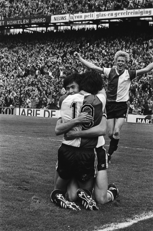 Drei Fußballspieler feiern ein Tor auf dem Boden vor einer jubelnden Menge in einem schwarz-weißen Stadion.