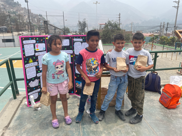 Eine Gruppe von Kindern steht auf einem Basketballfeld, jedes Kind hält eine Papiertüte in der Hand, mit Vorräten und einer Texttafel in der Nähe, und eine Kulisse aus Zäunen, Pfosten, Bäumen, Gebäuden und Hügeln.